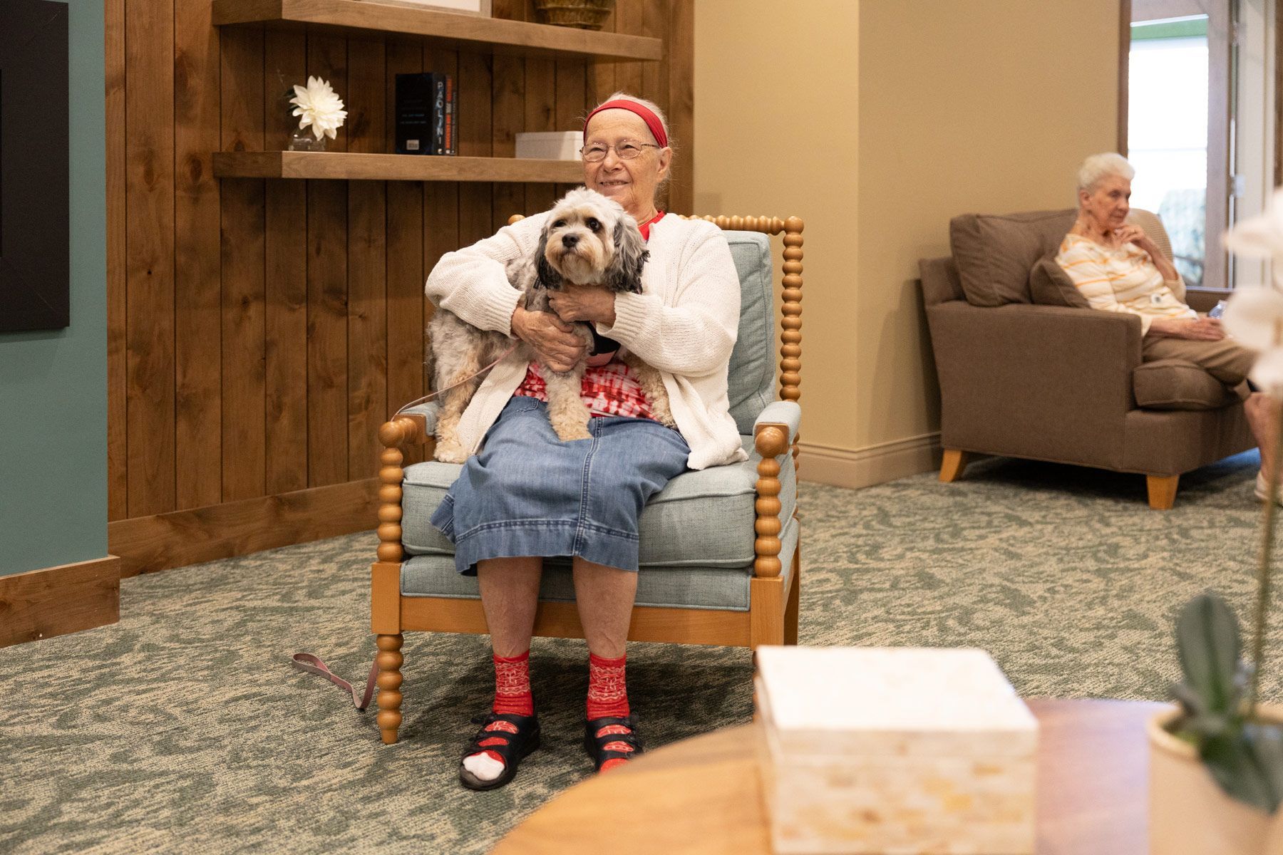 Woman sitting in chair, holding a dog, smiling. Another person sits in background. Interior room setting.
