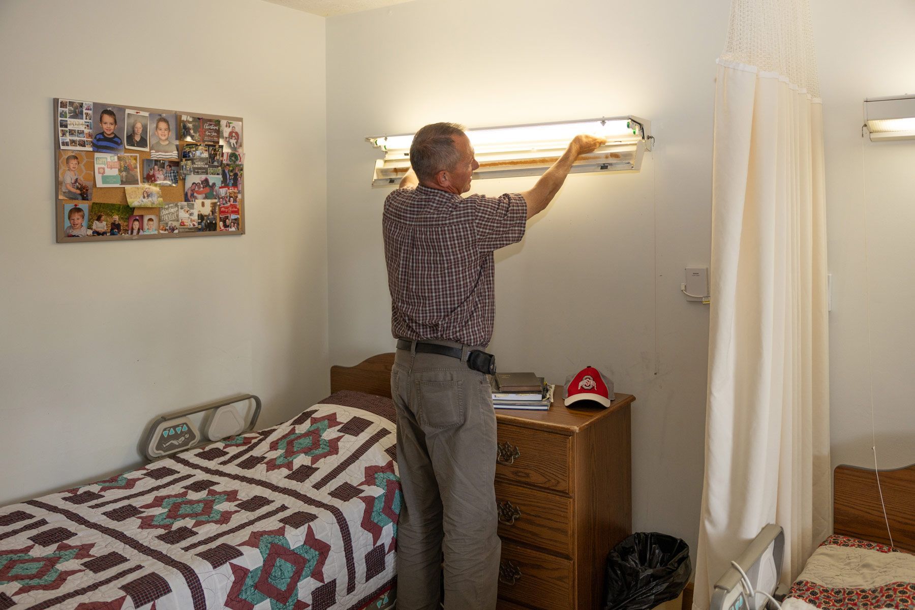 Man in a room, working on a light fixture above a wooden dresser. A bed is to the left.
