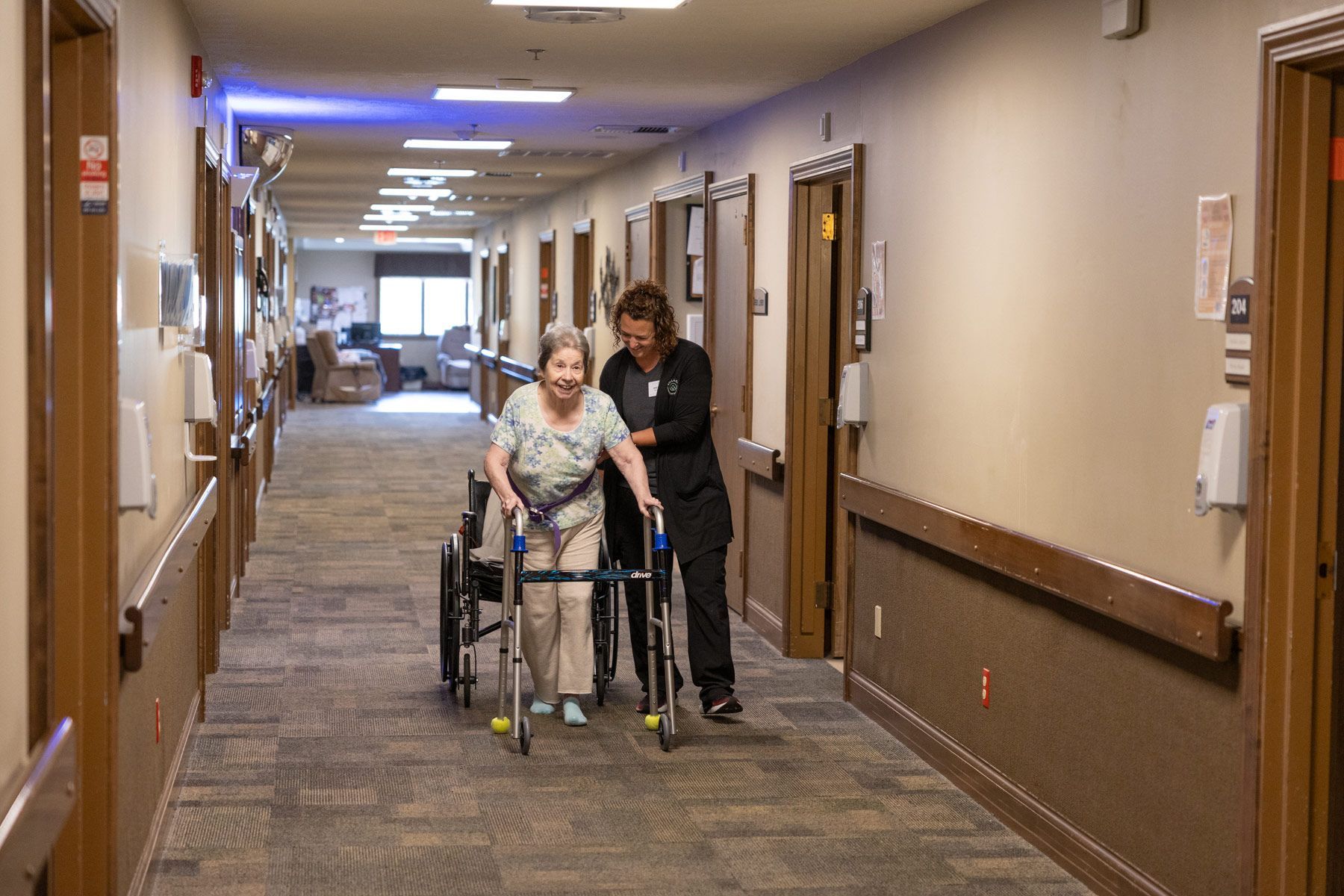 A healthcare worker assists a person using a walker down a long hallway with closed doors.