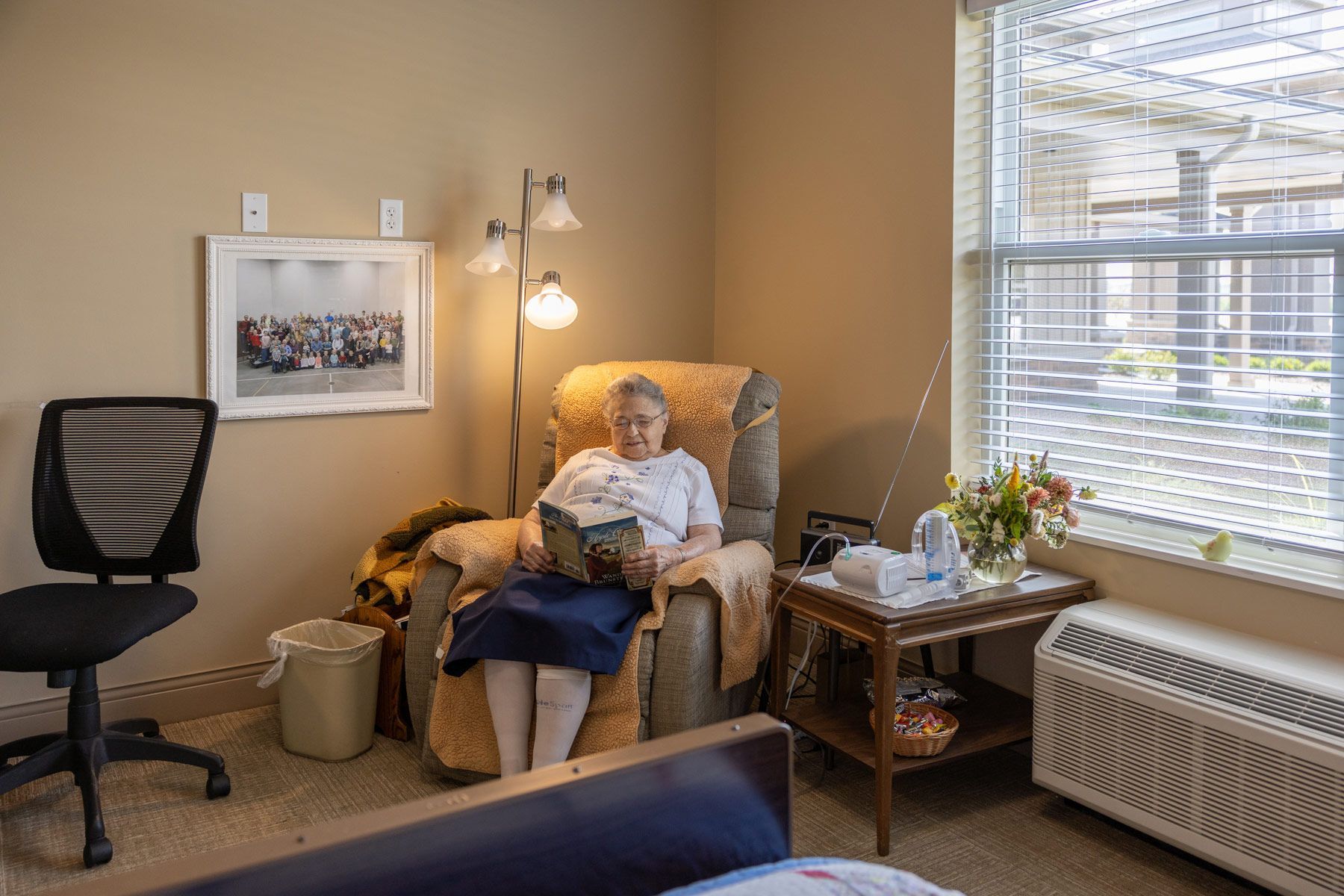 Woman reading in armchair by window, lamp, small table, artwork, and chair in a room.