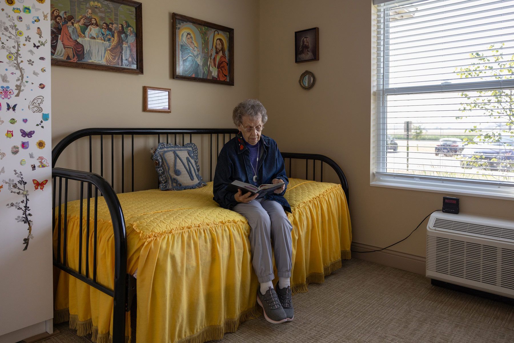 Woman sits on a bed, reading. She's in a room with a window and religious artwork on the wall.