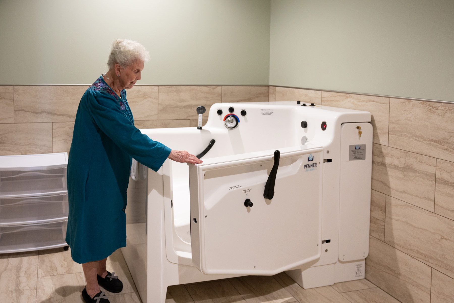Woman opening the door of a walk-in bathtub in a bathroom. She wears a blue robe.