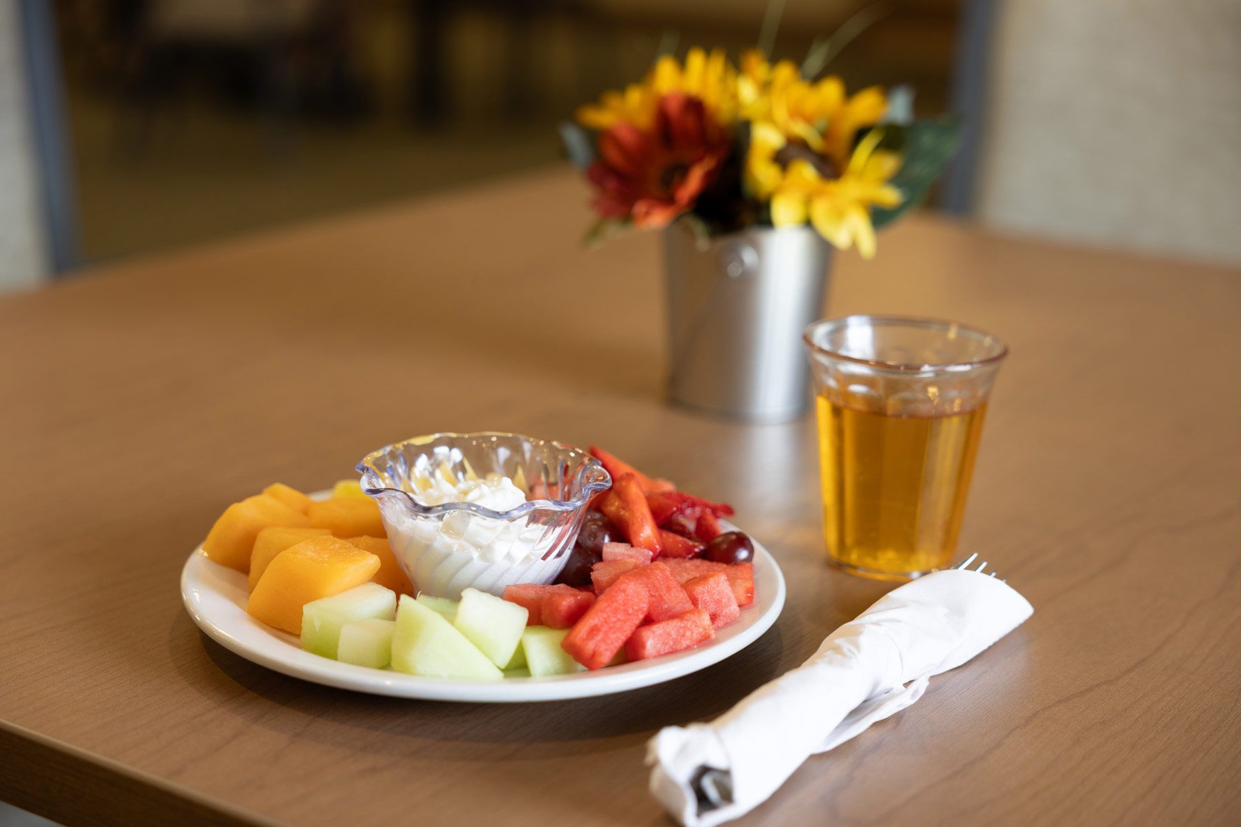 Plate of fruit with dip, glass of juice, and flowers on a table.
