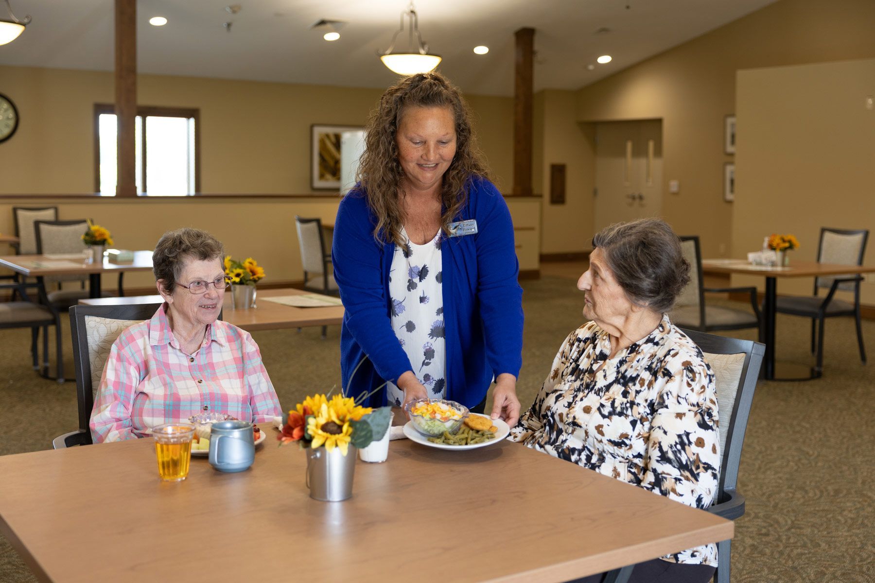 A caregiver serving a plate of food to a senior woman, with another woman seated at the table.