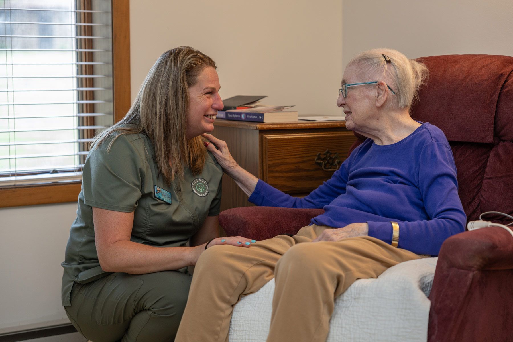 Caregiver comforts elderly person seated in chair; inside a home.