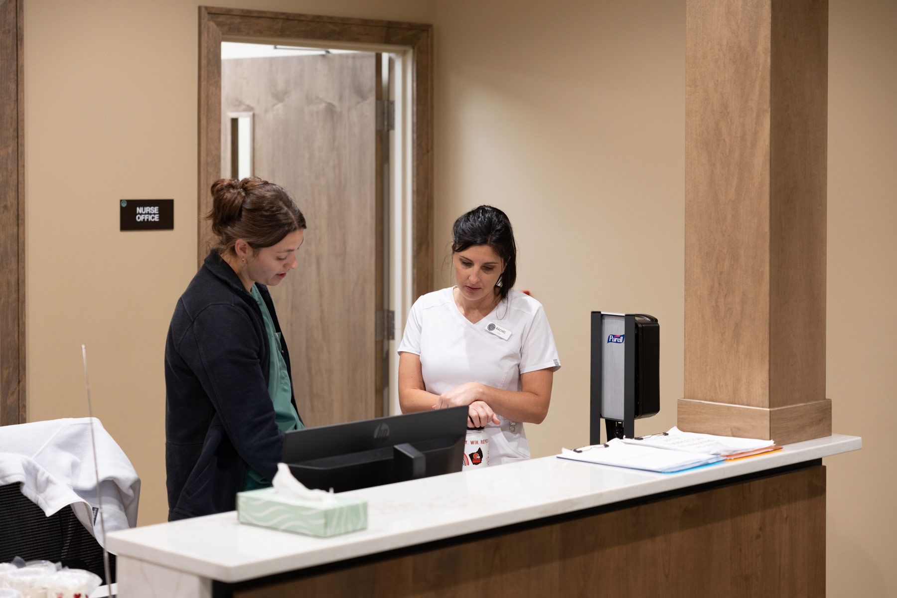 Two people at a reception desk, one looking at a computer, the other at a watch, in a clinic.