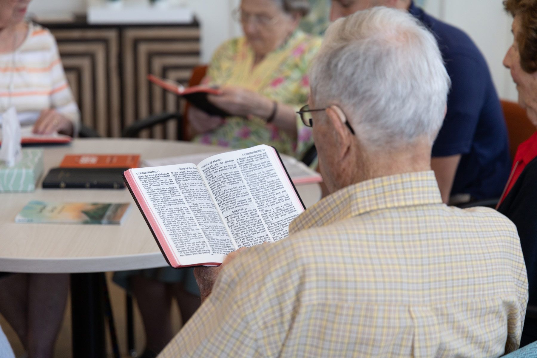 Group of people reading books around a table indoors. Focus on an elderly man with glasses reading.