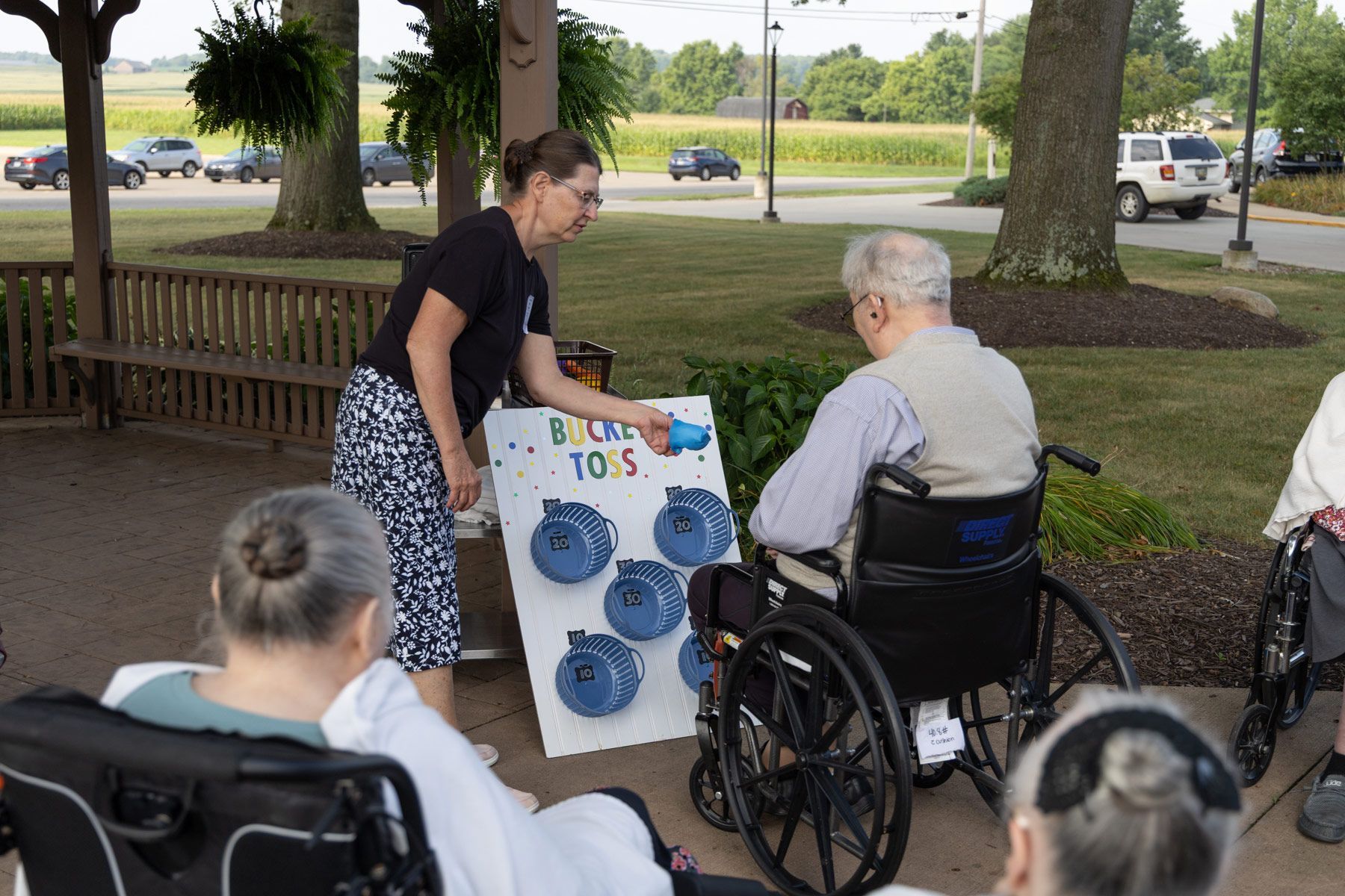 Woman helps senior in wheelchair play a beanbag toss game outdoors.