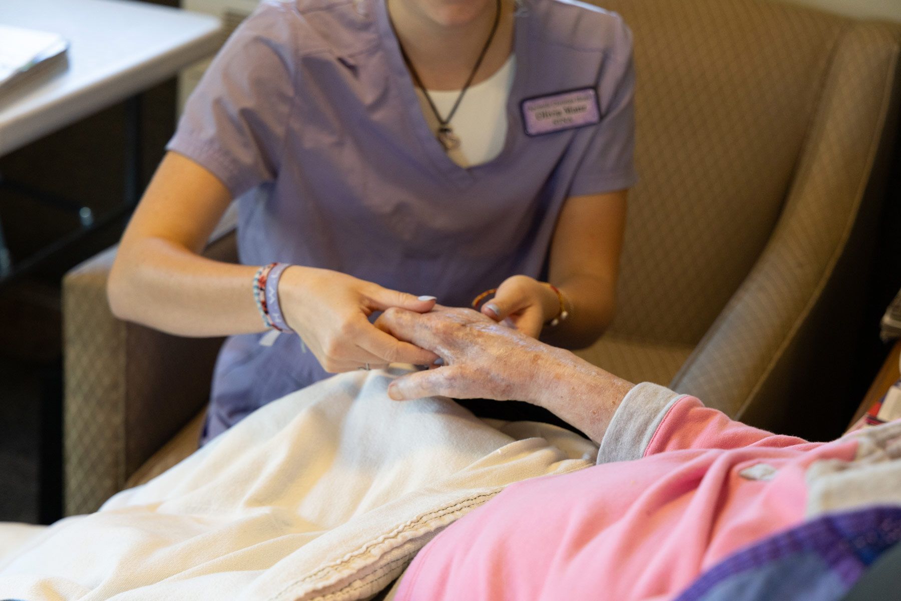 Person in scrubs gently massages the hand of a person lying in bed, likely in a care facility.
