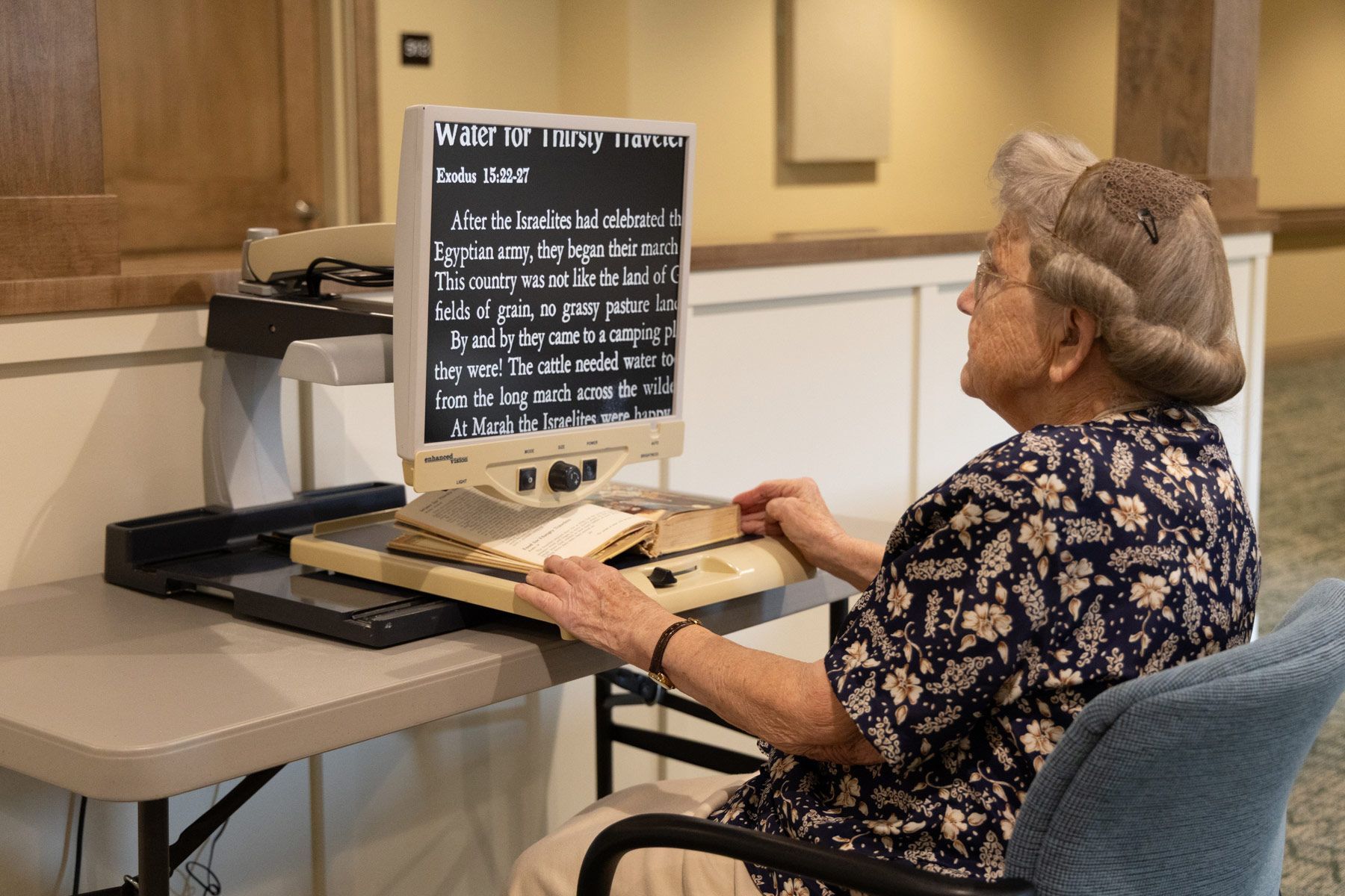 Woman using a desktop video magnifier to read a book, seated at a table.