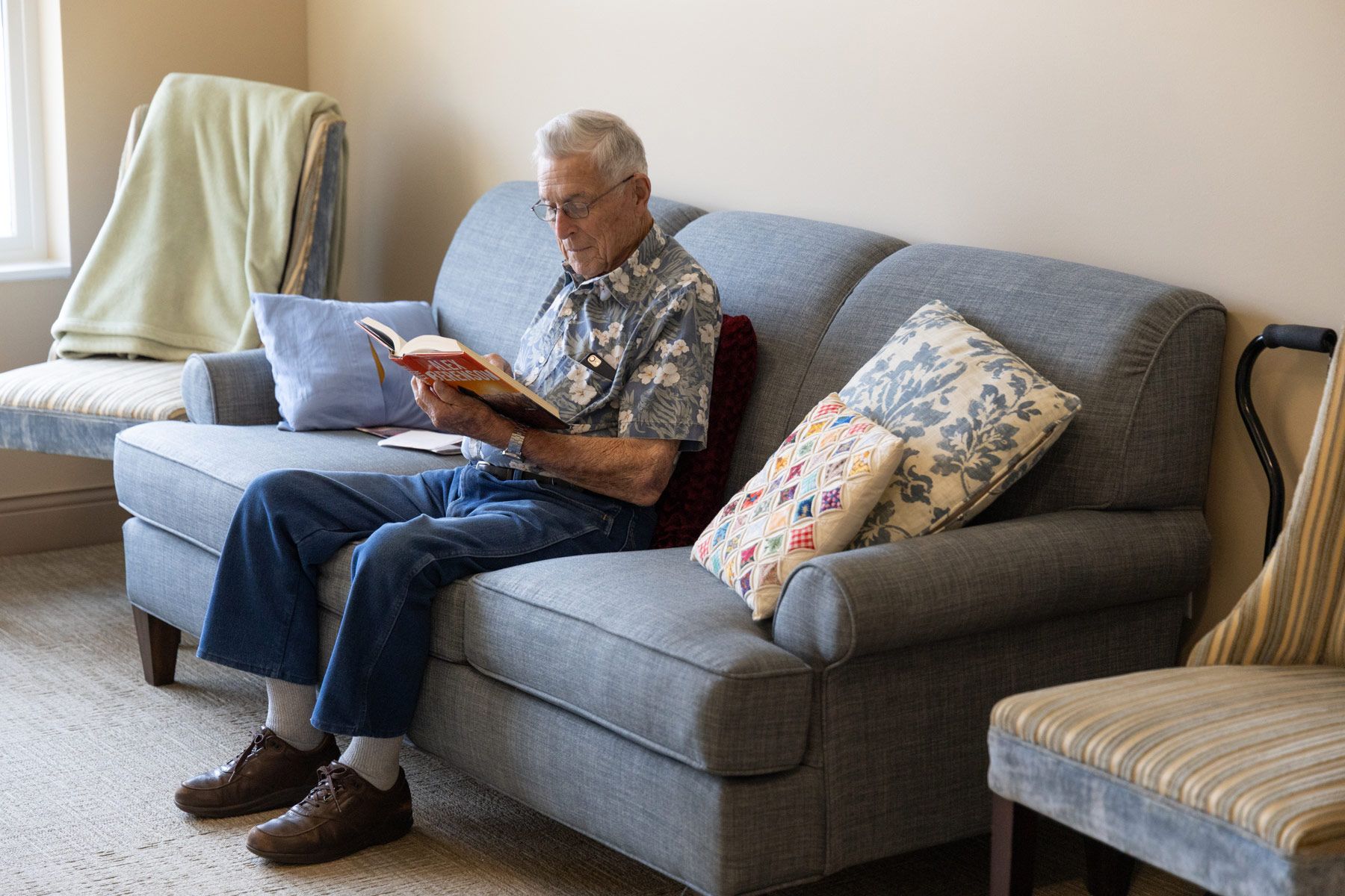 Older person reading a book on a gray couch with pillows, indoors.