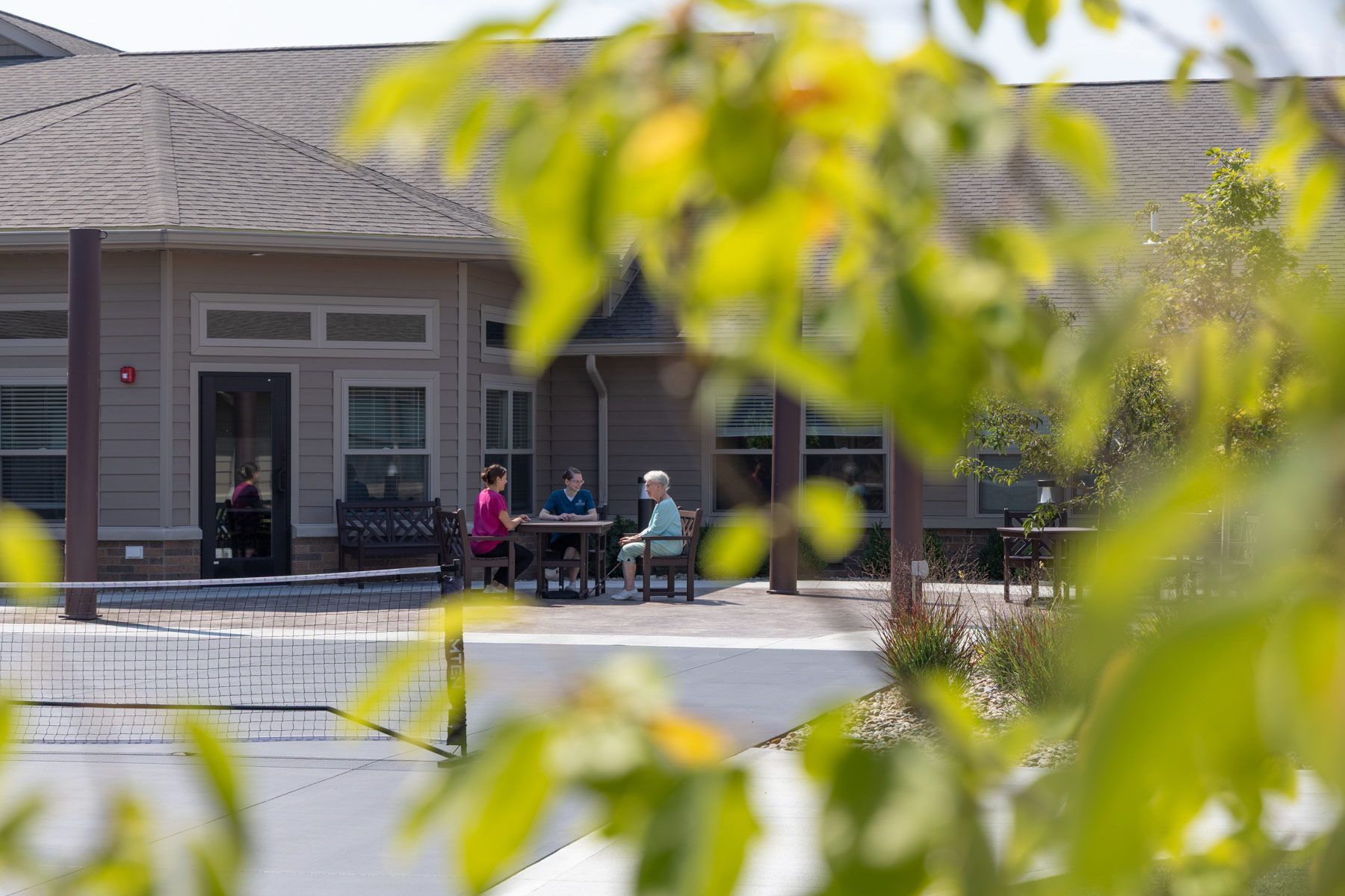 Three people sit at an outdoor table on a patio. Tan building with dark roof in background, greenery in foreground.
