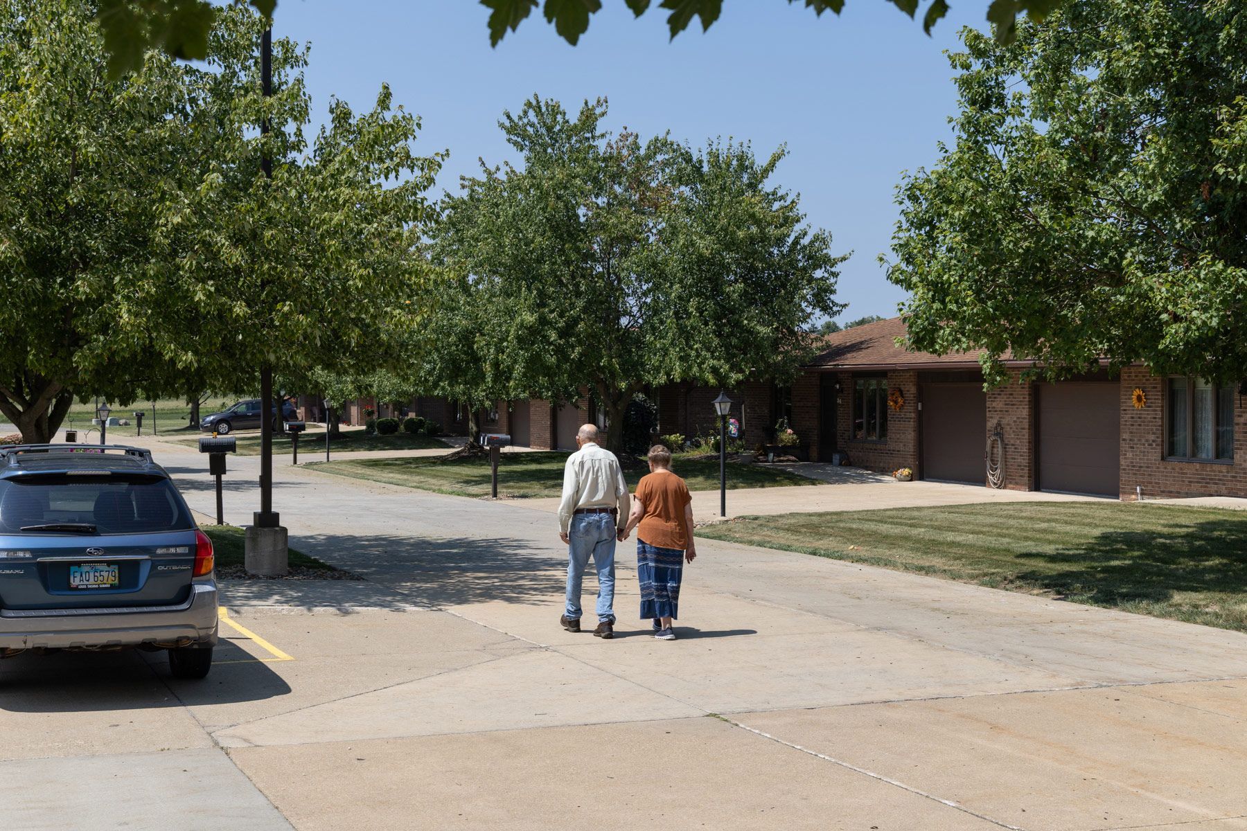 Couple walking hand-in-hand on a paved path in front of a row of brick buildings and trees on a sunny day.