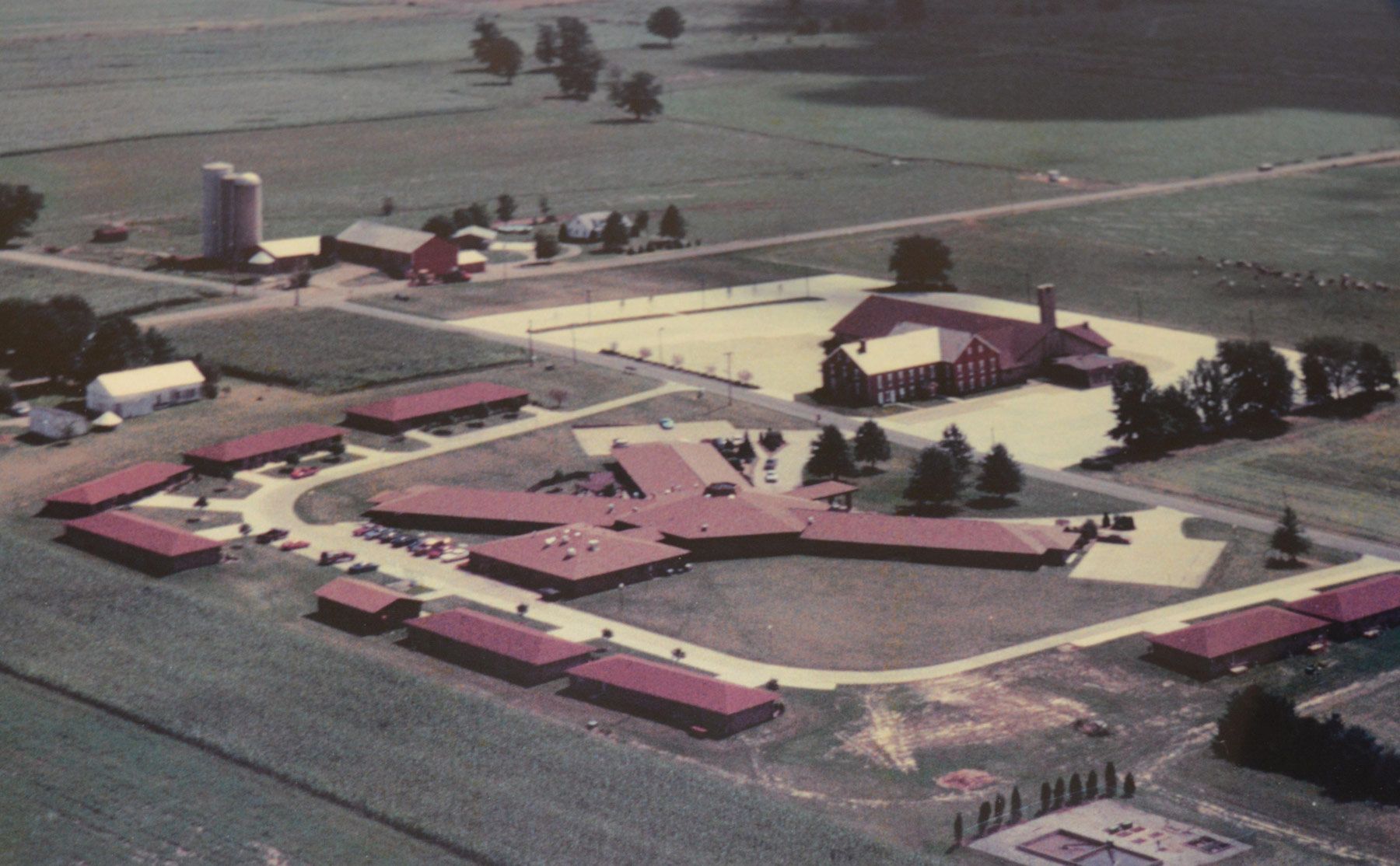 Aerial view of a complex with long, low buildings and a central structure, likely a facility or campus in a rural setting.