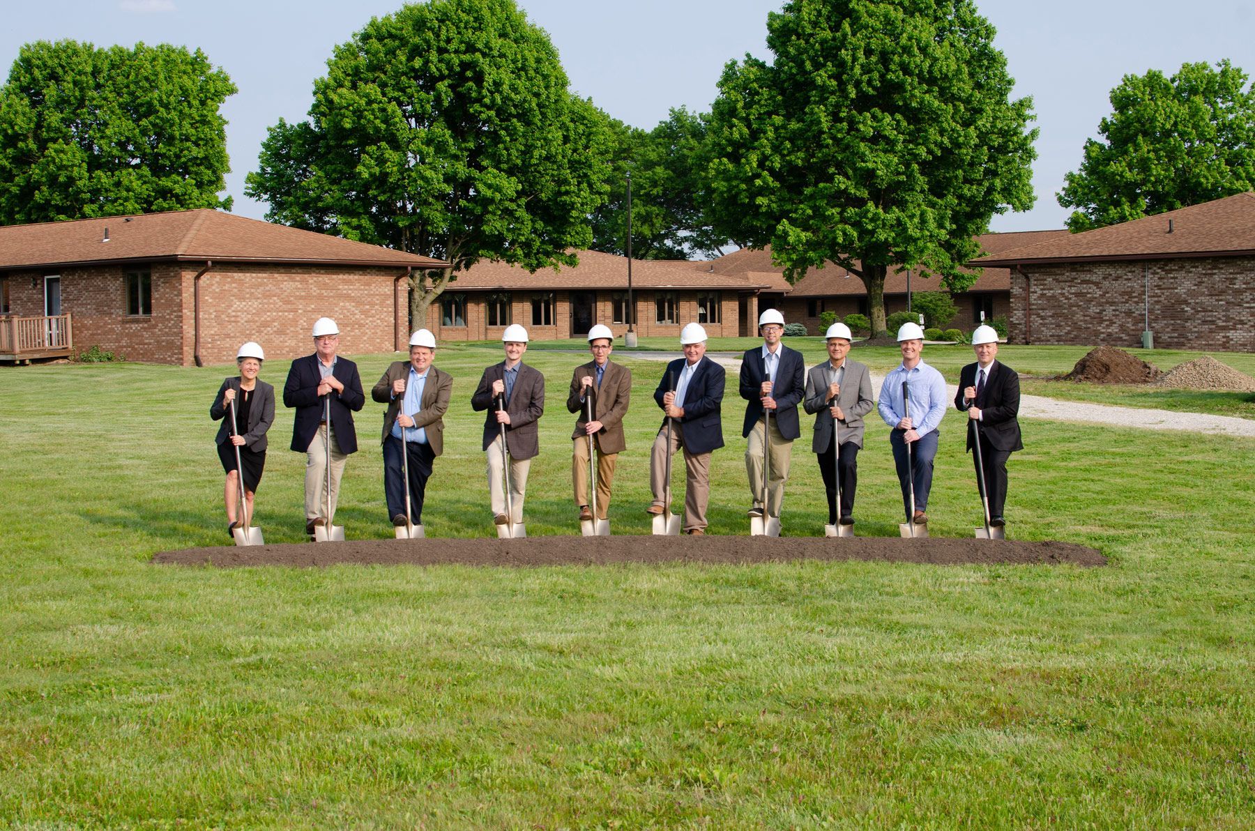 Group of people with shovels at a groundbreaking ceremony, in front of a brick building.