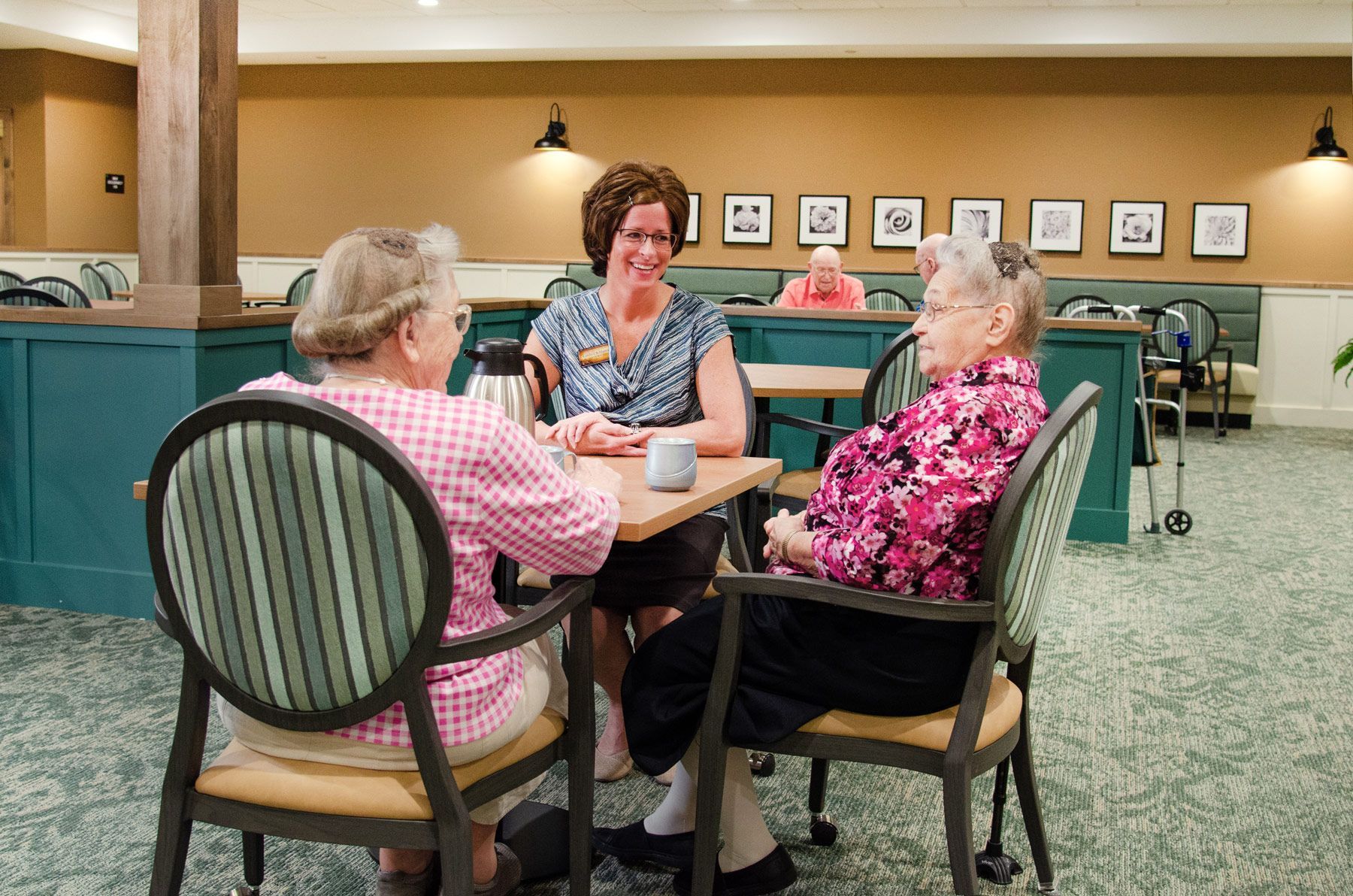 Three people seated at a table; two elderly women and a smiling staff member. They are in a dining room.