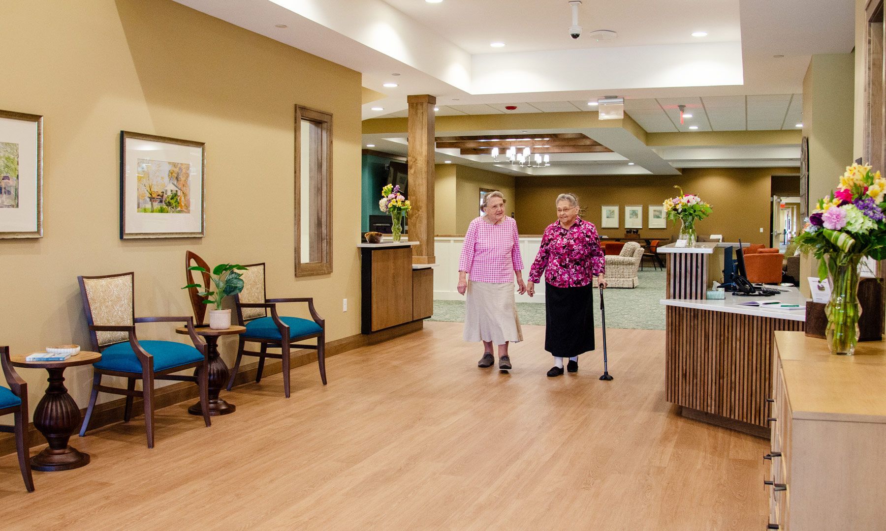 Two older women walking in a senior living facility, one using a cane, near a reception desk.