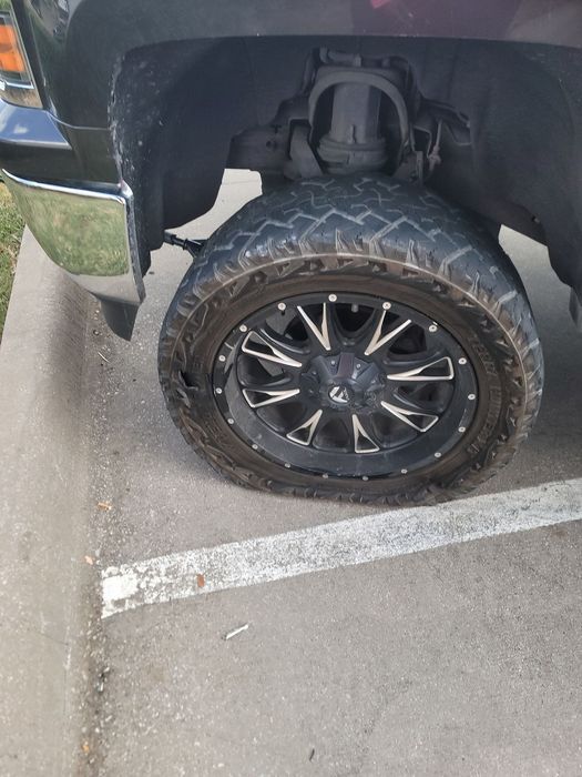 A close-up view of a truck's front tire, showing a flat, damaged tire mounted on a black and silver aftermarket rim.