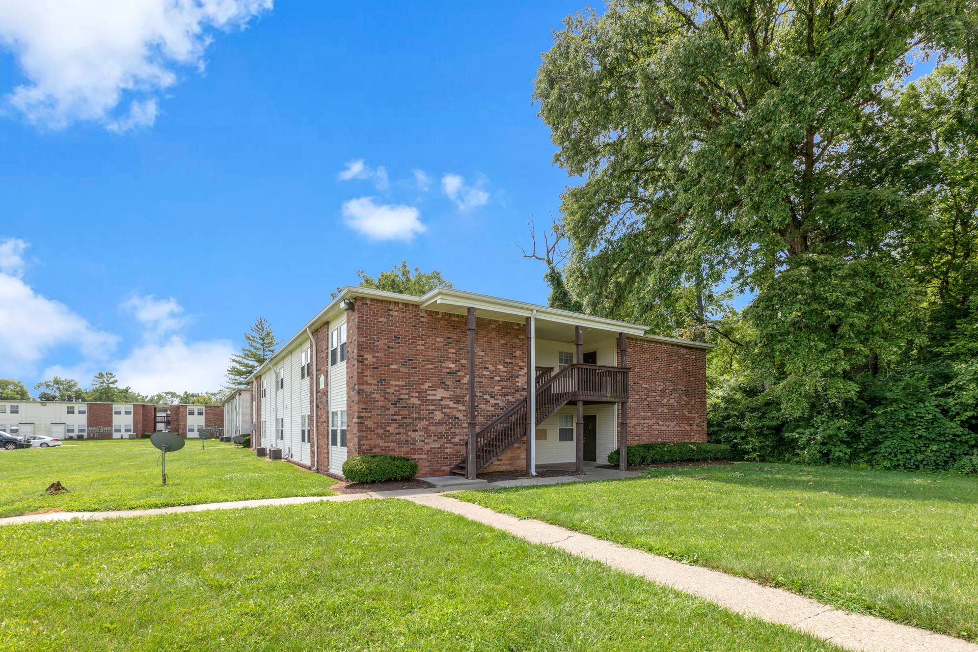 Two-story brick apartment building with a wooden stairwell, surrounded by green grass and trees, under a blue sky.