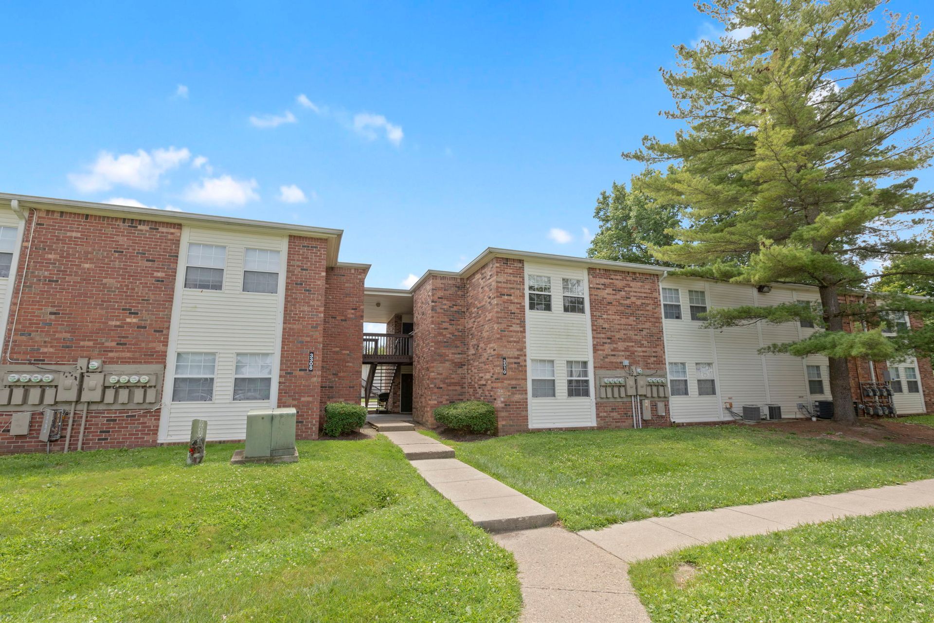 Two-story brick and white apartment buildings with green lawns and a concrete walkway under a blue sky.
