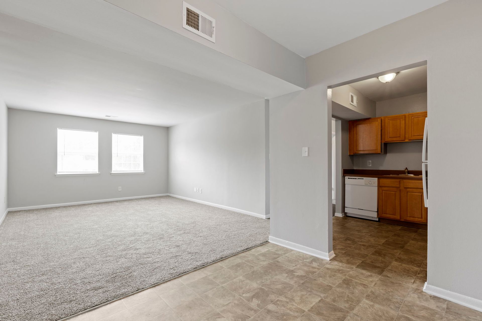 Empty apartment interior with carpet, windows, and a view into the kitchen.
