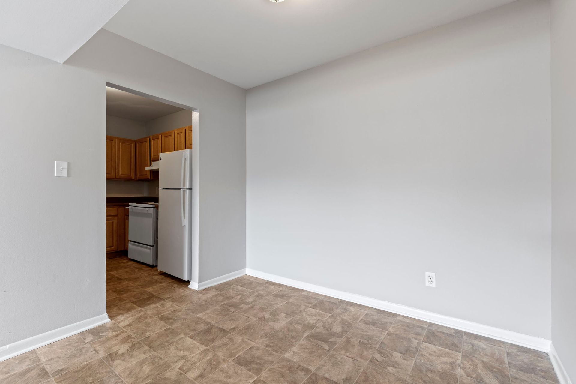 Empty room with light gray walls, brown floor, and a doorway leading to a kitchen.