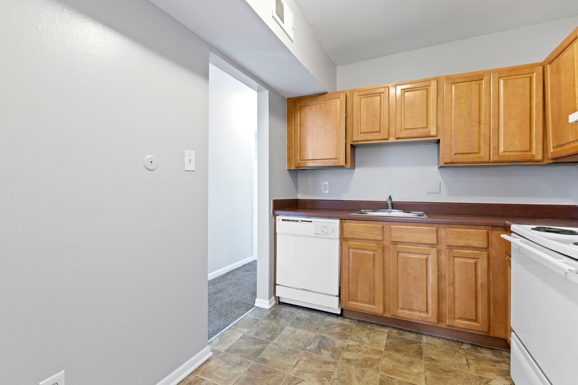 Kitchen with wooden cabinets, white appliances, and gray walls; doorway leads to a carpeted hallway.