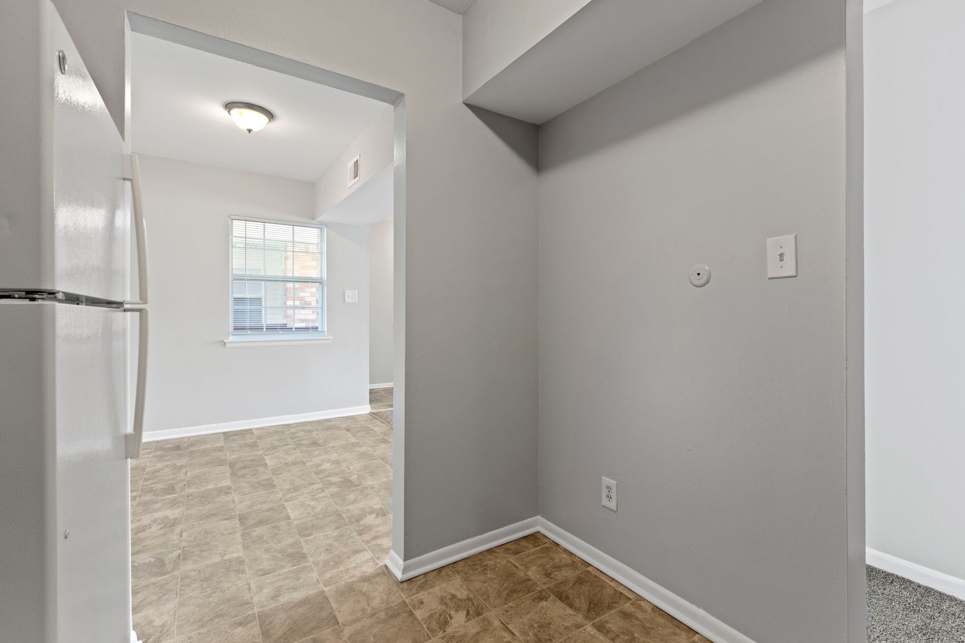 Interior shot of an apartment kitchen and doorway leading to another room. Gray walls, white fridge, and tan flooring.