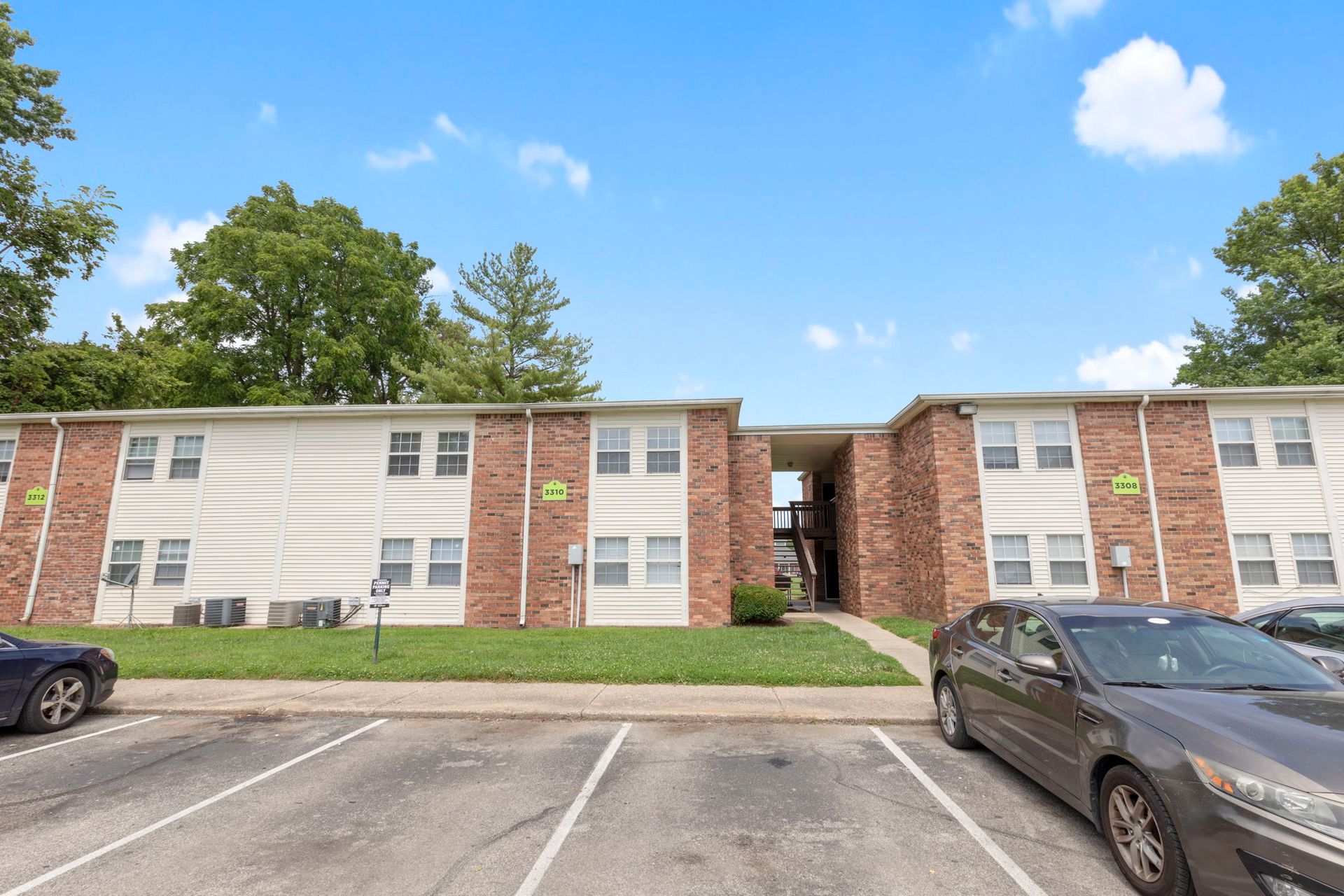 Apartment building with brick and white siding, cars in parking spots, green grass, blue sky.