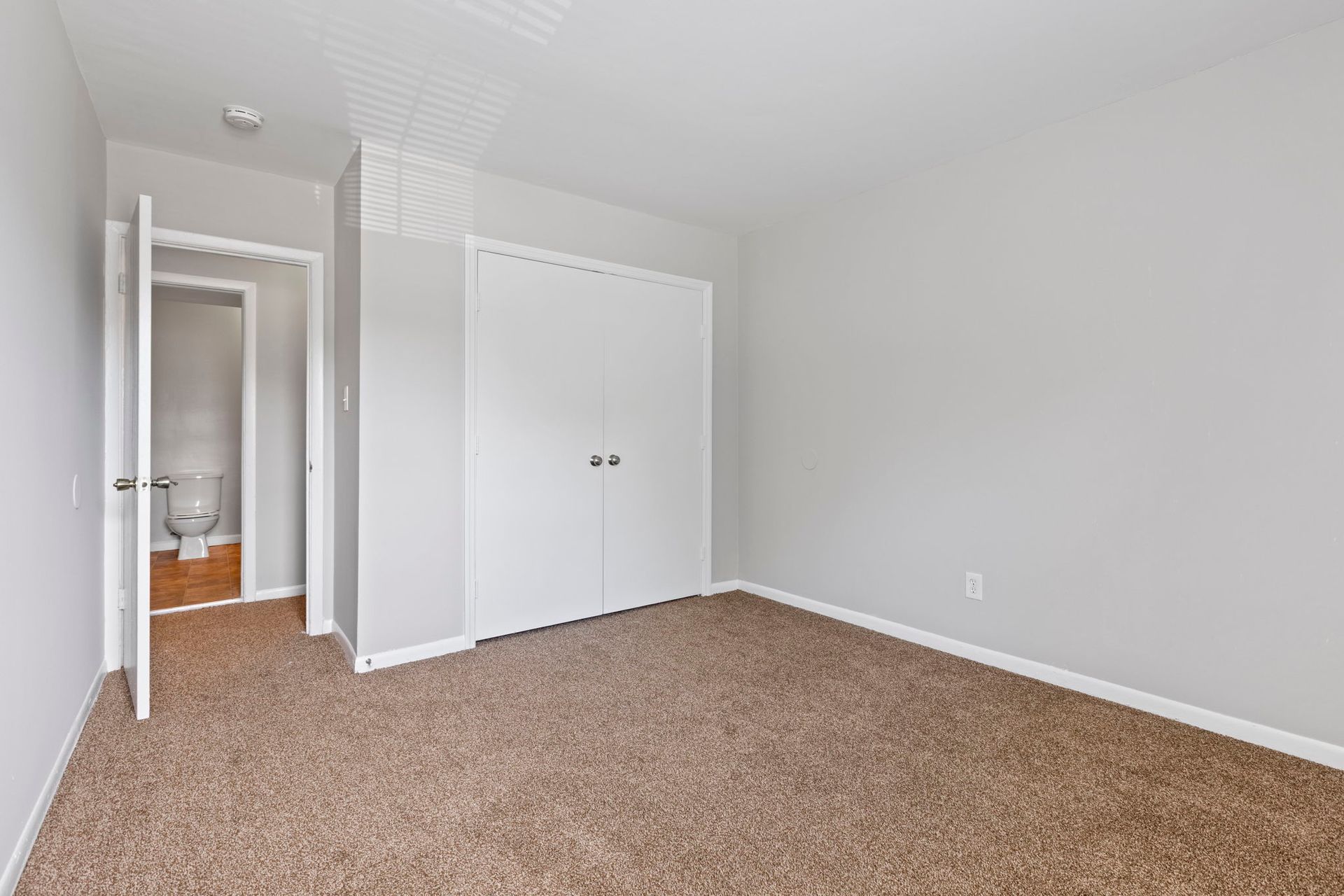 Empty bedroom with brown carpet, gray walls, white closet doors, and open bathroom door.