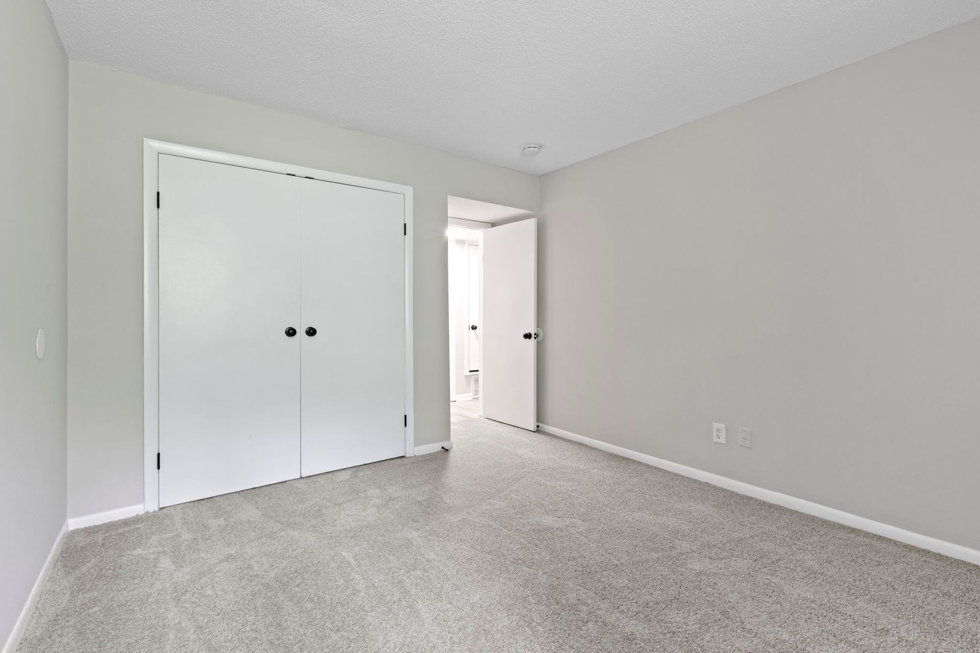 Empty bedroom with gray carpet, white closet doors, and a doorway to a bathroom.