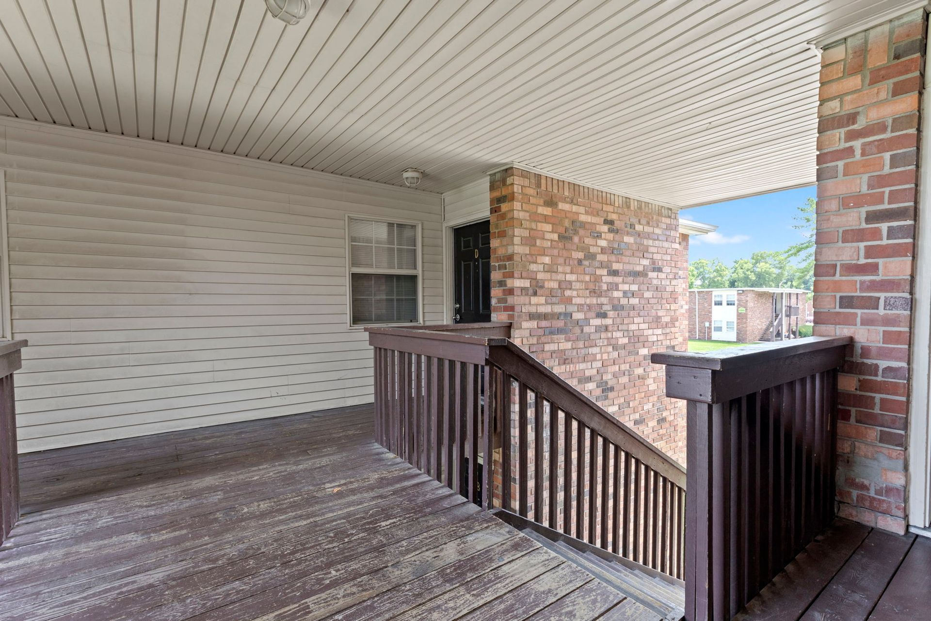 Covered porch with wooden deck, railing, brick wall, and entry door.