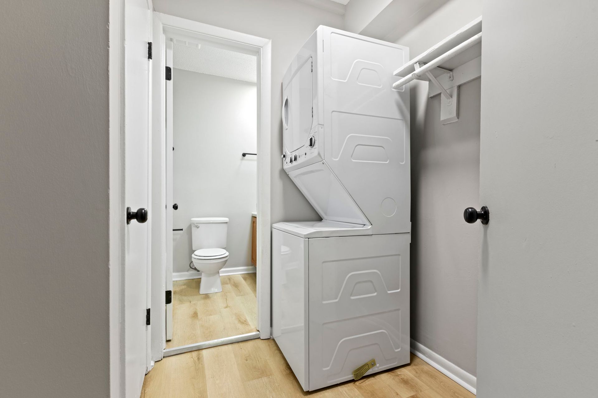 A small laundry room with a stacked washer and dryer, toilet visible through open doorway.