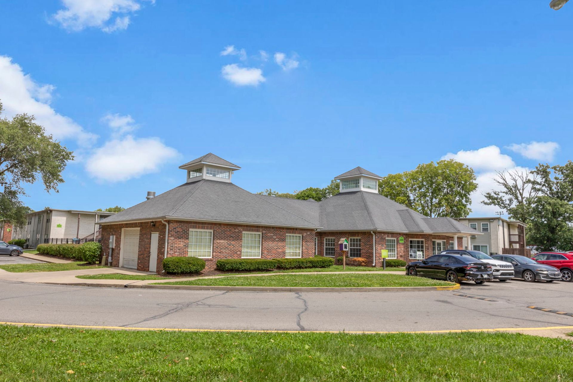 Brick building with two cupolas, cars parked in front, and a blue sky.