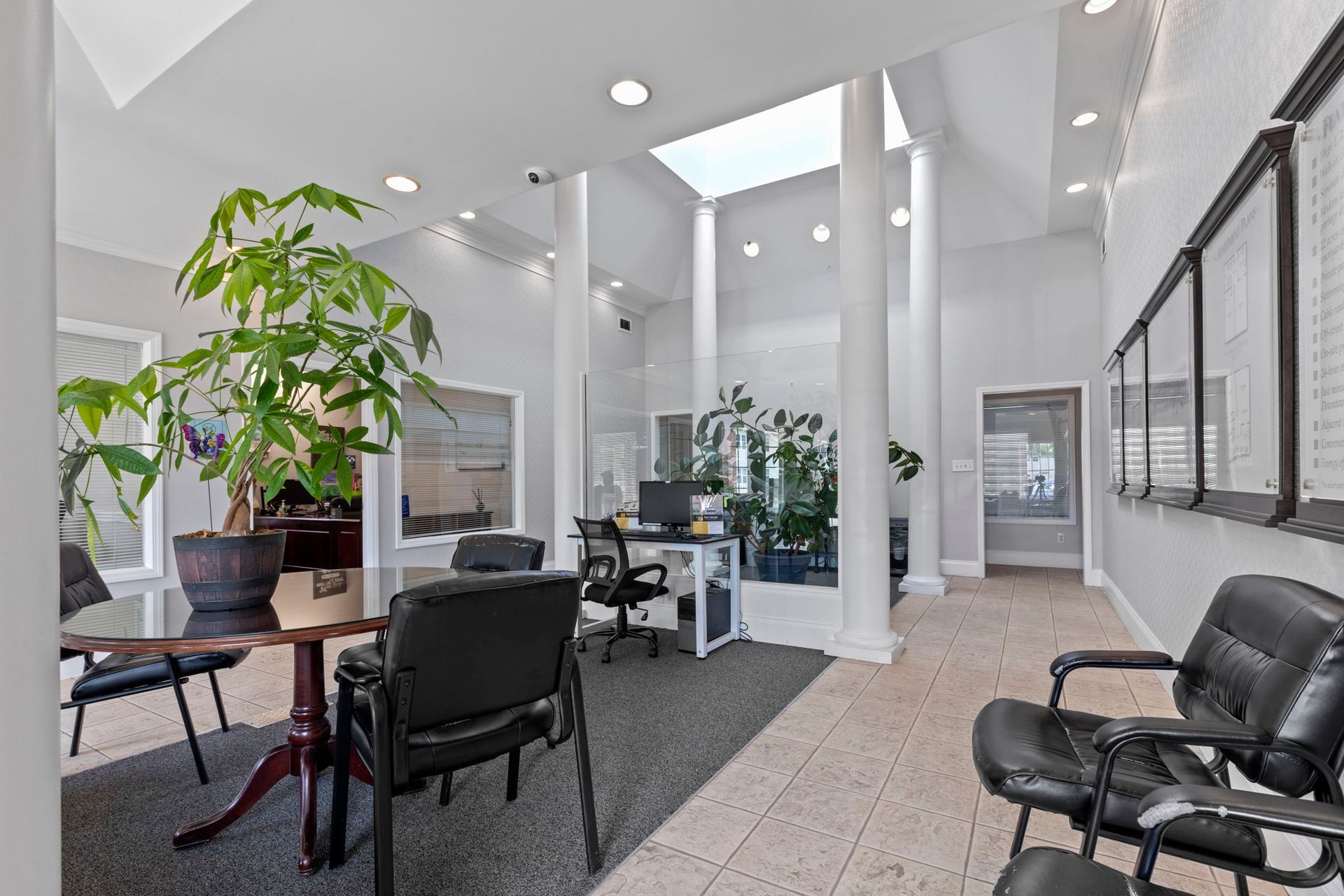 Reception area with round table, black chairs, potted plant, and a desk. White walls, skylight.