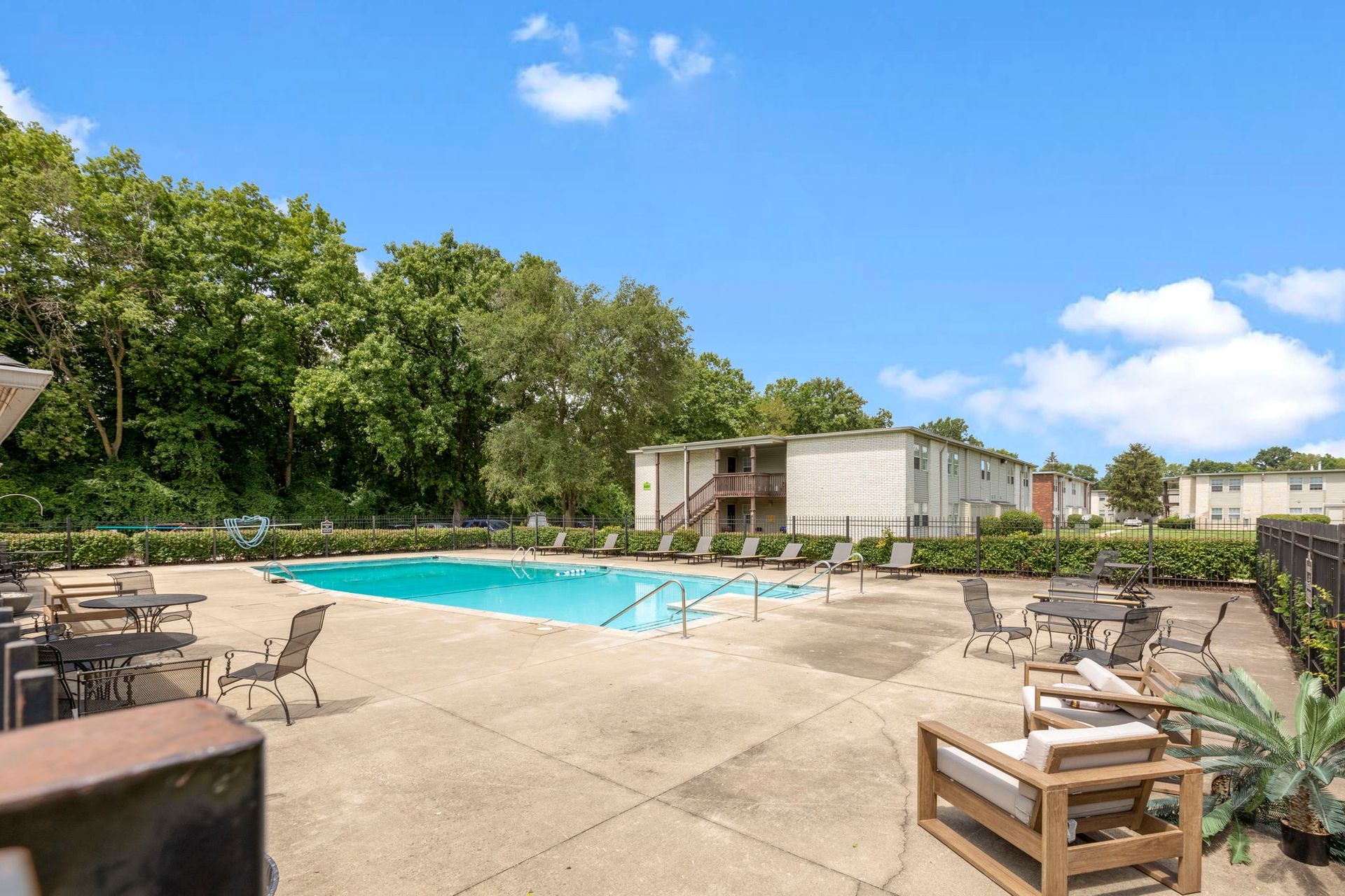 Pool area with patio furniture, apartment building, trees, and blue sky.