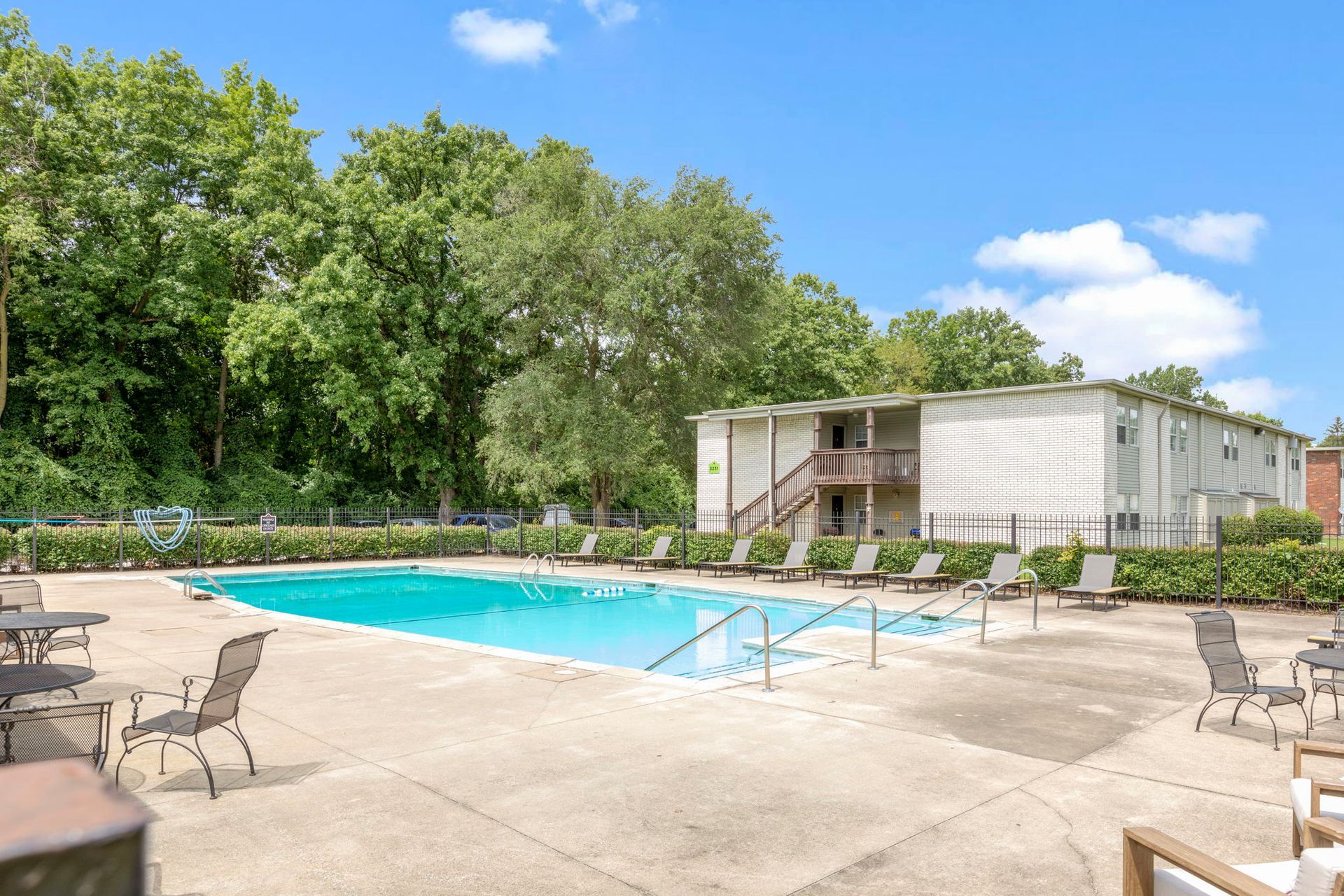 Apartment complex with pool, lounge chairs, and lush trees on a sunny day.