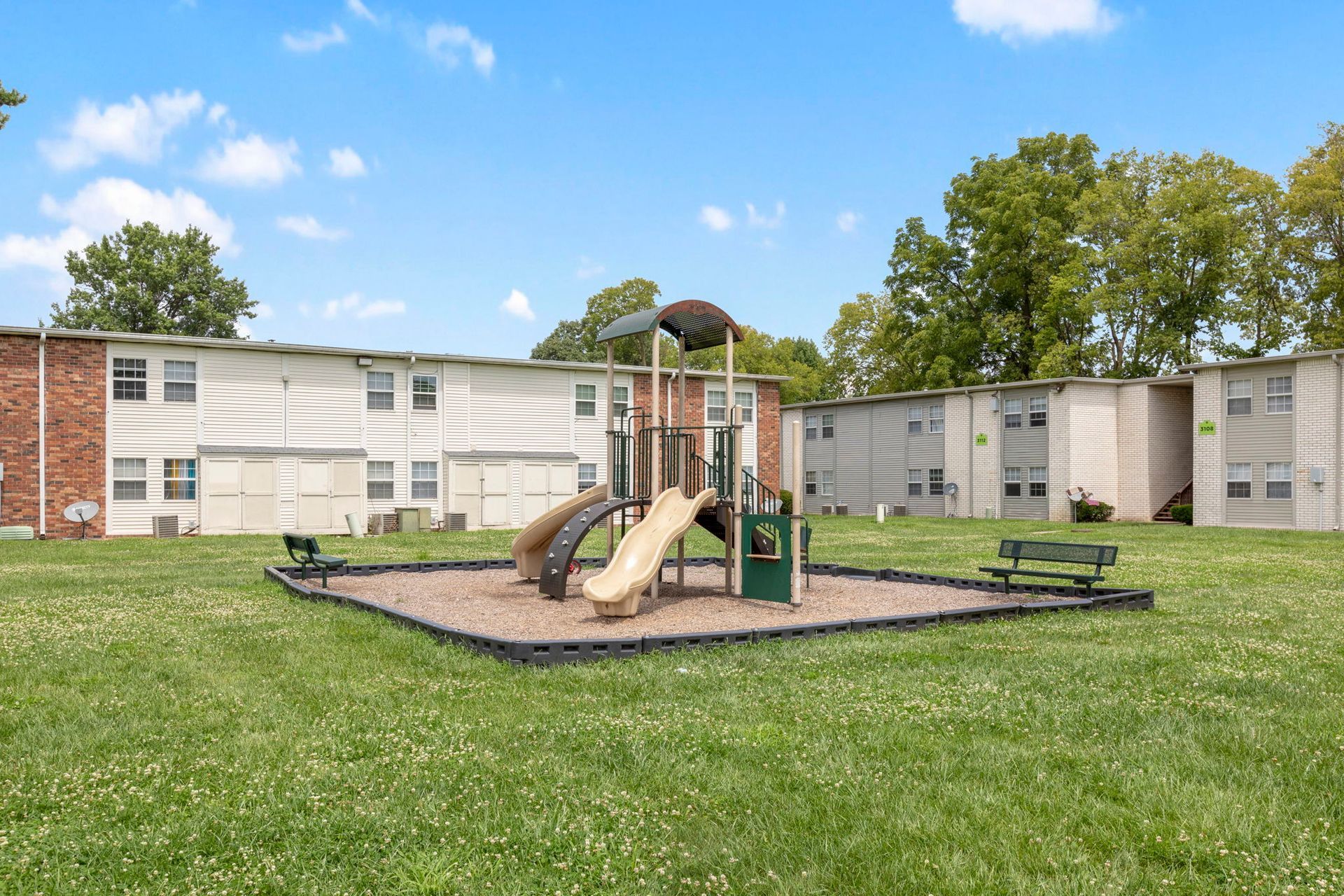 Playground in front of apartment buildings, with slides, climbers, and benches, on a grassy lawn under a blue sky.