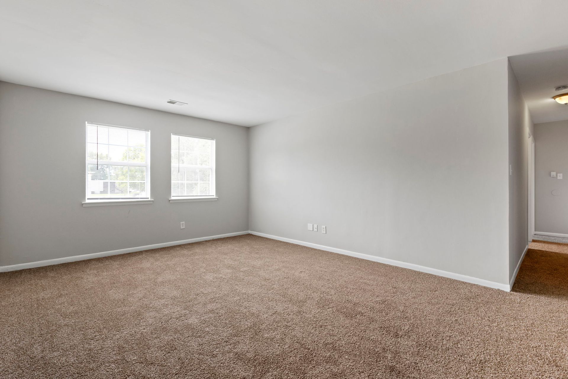 Empty room with beige carpet, two windows, and gray walls. Hallway on the right.