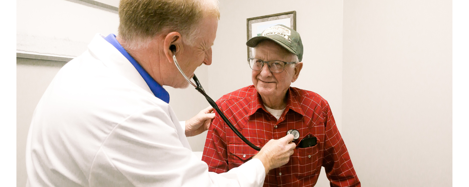 Doctor using a stethoscope to examine a smiling patient in a red shirt and cap.