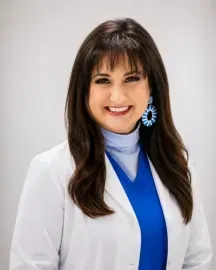 Smiling woman in a white blazer with blue top and earrings, against a light background