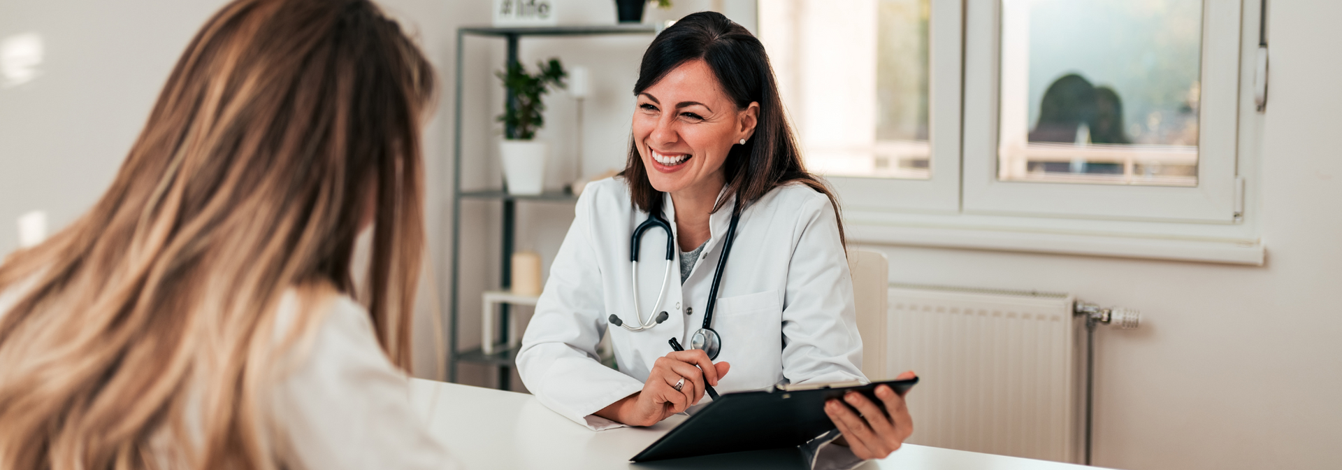 Doctor speaking with a patient in a bright clinic office, holding a tablet and smiling.