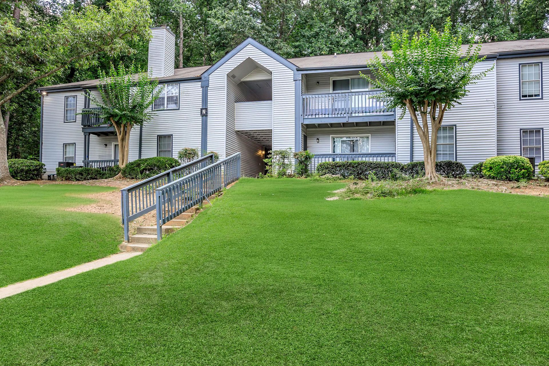 Ranch-style house with brick and gray exterior, pillars, surrounded by greenery and lawn.