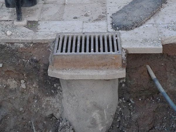 Drainage grate in a concrete surround, installed in a dirt excavation, with paving stones nearby.