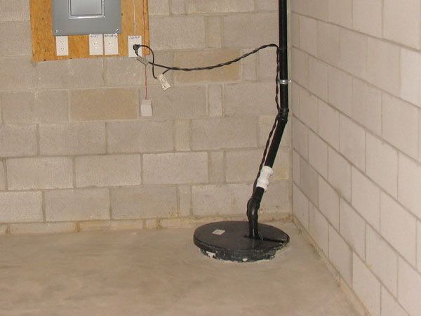 Sump pump in basement corner with electrical panel in the background and cinder block walls.