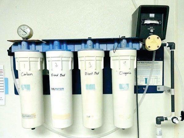 Water filtration system with four white filter cartridges, gauges, and tubes mounted on a wall.
