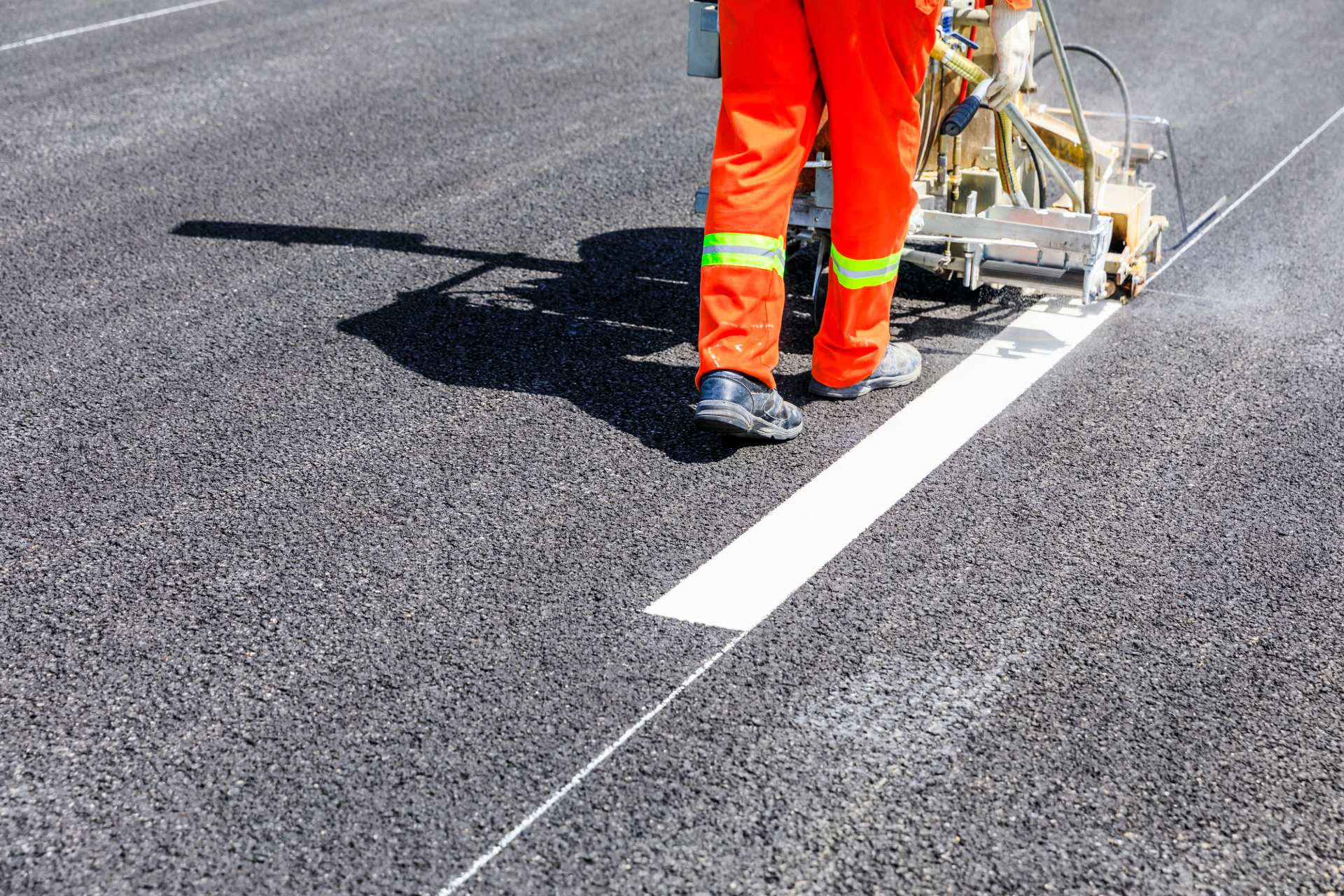 Road worker in orange pants painting a white lane line on asphalt with a road-marking machine.