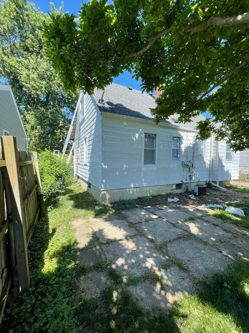 Backyard view of a white house with a cracked concrete patio and overgrown grass.