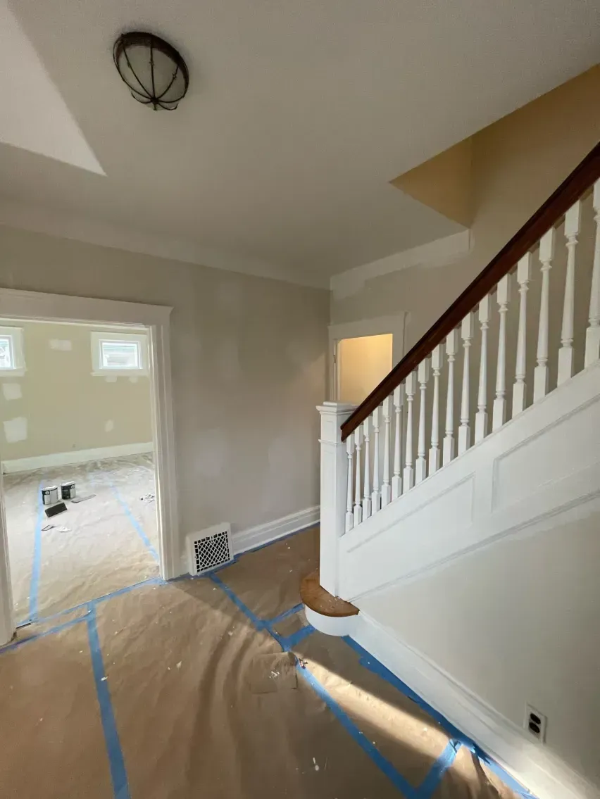 Interior of a home under renovation, hallway with staircase, doorway, and paper covering floors with blue tape.
