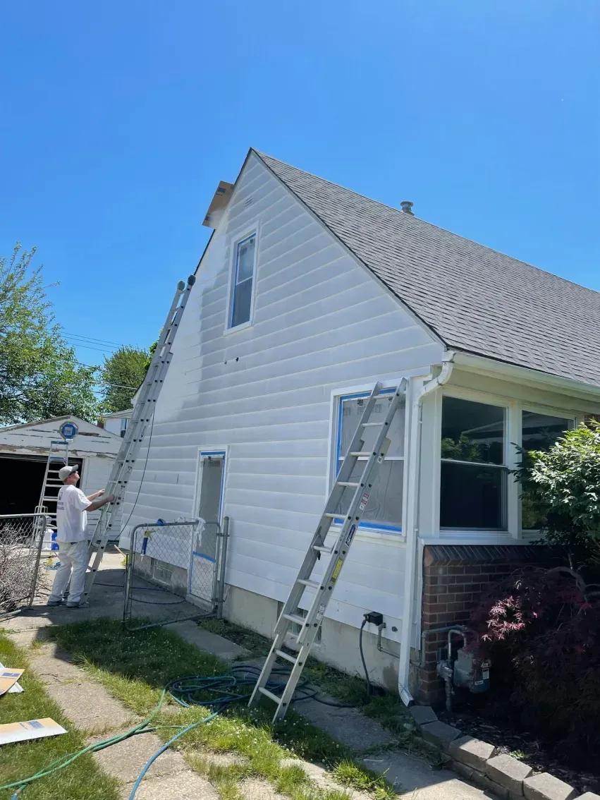 Person painting the side of a white house, using ladders. Sunny day, blue sky.